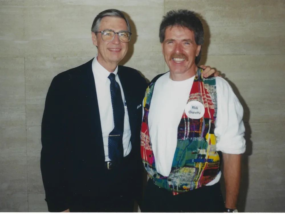 Fred Rogers, in a dark suit and glasses, with his arm around Rick Charette, who wears a patchwork vest and a conference name badge.
